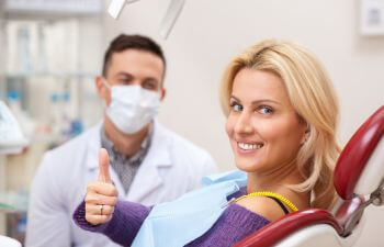 A dentist and a satisfied mature woman with a perfect smile sitting in a dental chair and showing her thumb up.
